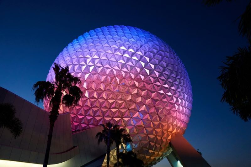 The image shows Spaceship Earth at Epcot, a geodesic sphere illuminated with a display of blue, pink, and orange lights against the dark night sky. Palm trees are visible in the foreground, framing the structure.