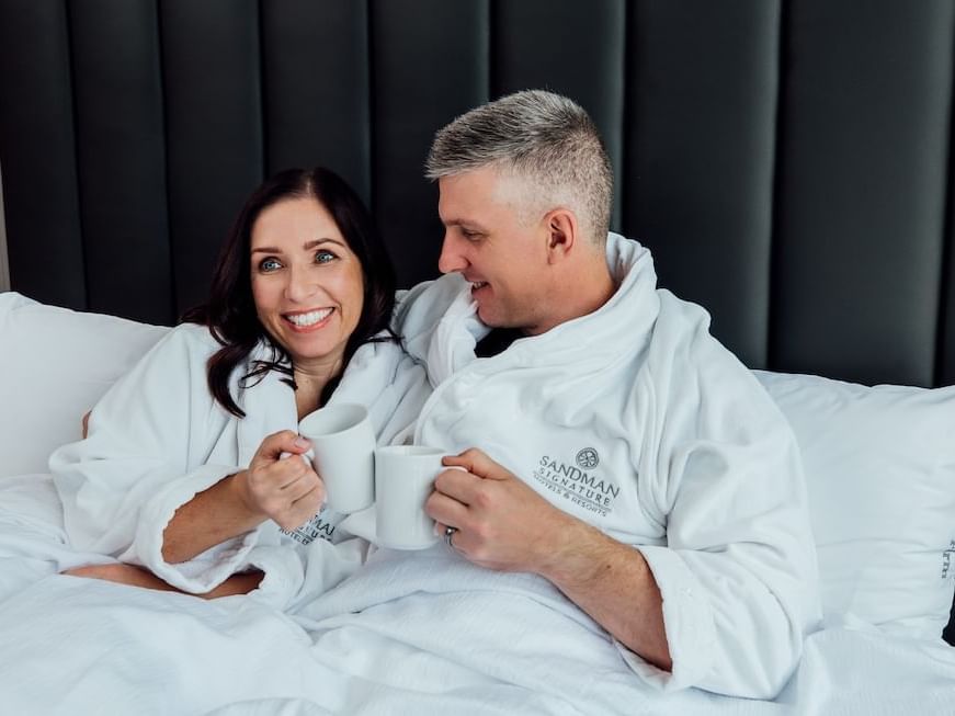 Two people in white robes holding coffee mugs in bed at Sandman Hotel & Suites Winnipeg Airport