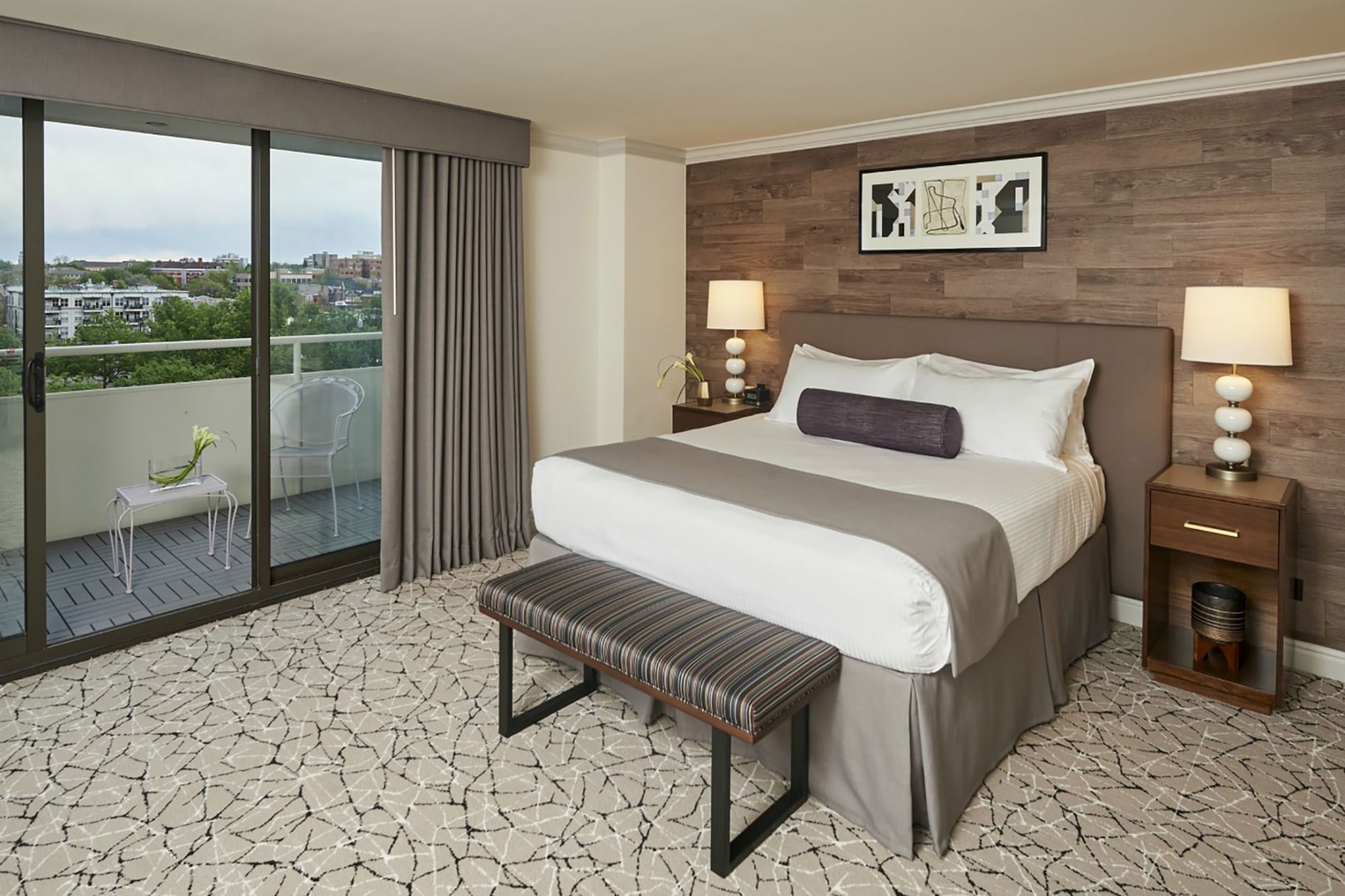 A hotel room with a bed, nightstands, a bench, and a balcony with a city view.