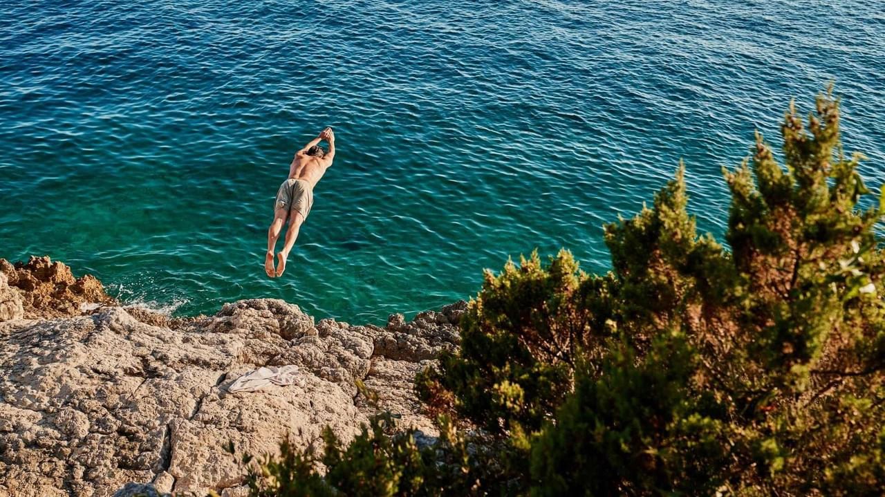 Mann springt in klares blaues Wasser von einem felsigen Ufer mit grünen Pflanzen.