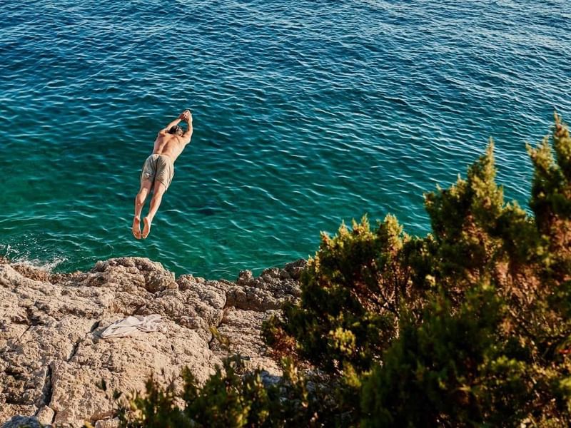 Mann springt in klares blaues Wasser von einem felsigen Ufer mit grünen Pflanzen.
