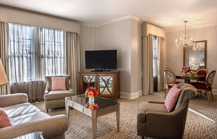 Living area with a TV on a wood cabinet by armchairs under a crystal chandelier in Deluxe Suite at Warwick Melrose Dallas