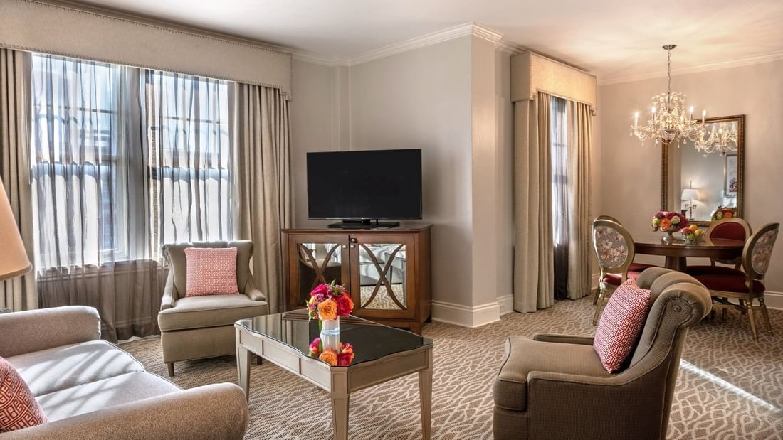 Living area with a TV on a wood cabinet by armchairs under a crystal chandelier in Deluxe Suite at Warwick Melrose Dallas