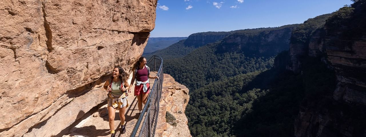 Two women hike on a cliffside path with railings, overlooking a valley and forest.