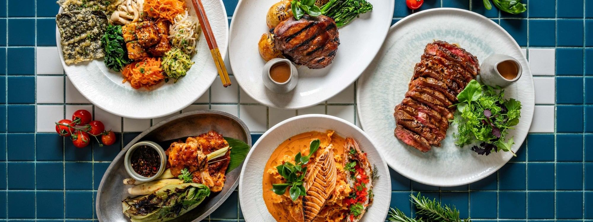 Overhead view of five gourmet dishes including grilled meats, seafood, and vegetables arranged on white plates over blue tiles.