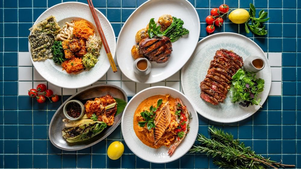 Overhead view of five gourmet dishes including grilled meats, seafood, and vegetables arranged on white plates over blue tiles.
