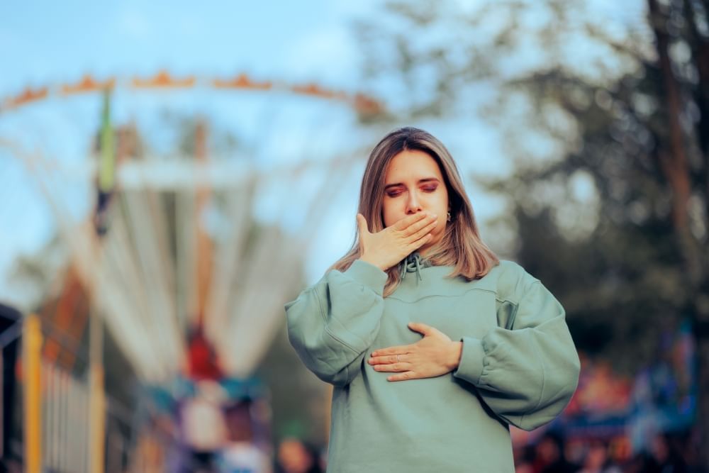 A nauseous-looking woman holds her mouth and her stomach with out of focus carnival rides in the background.