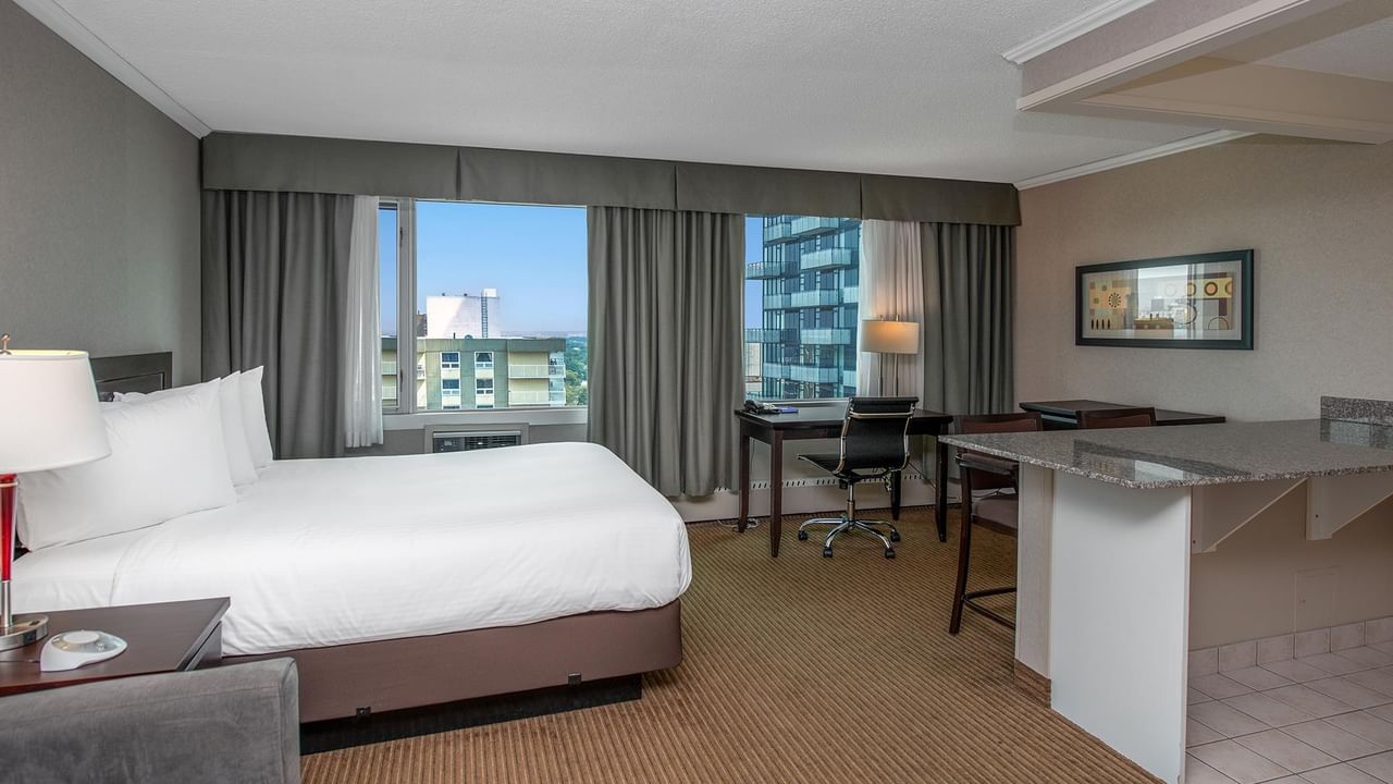 Hotel suite with a queen bed, work desk, kitchenette and large windows.