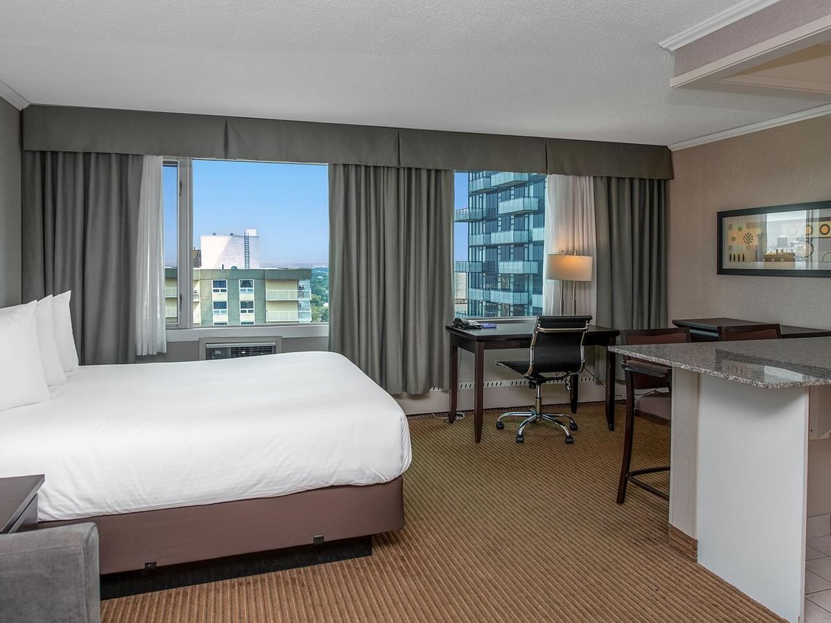Hotel guest room with a queen bed, work desk, kitchenette and large windows.