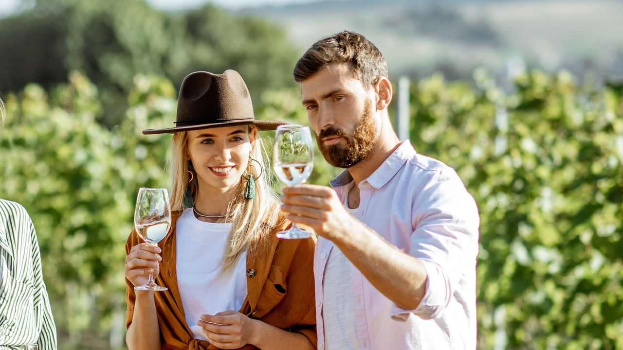 Couple holding glasses and enjoying wine in a vineyard, surrounded by lush green grapevines near Coast Penticton Hotel