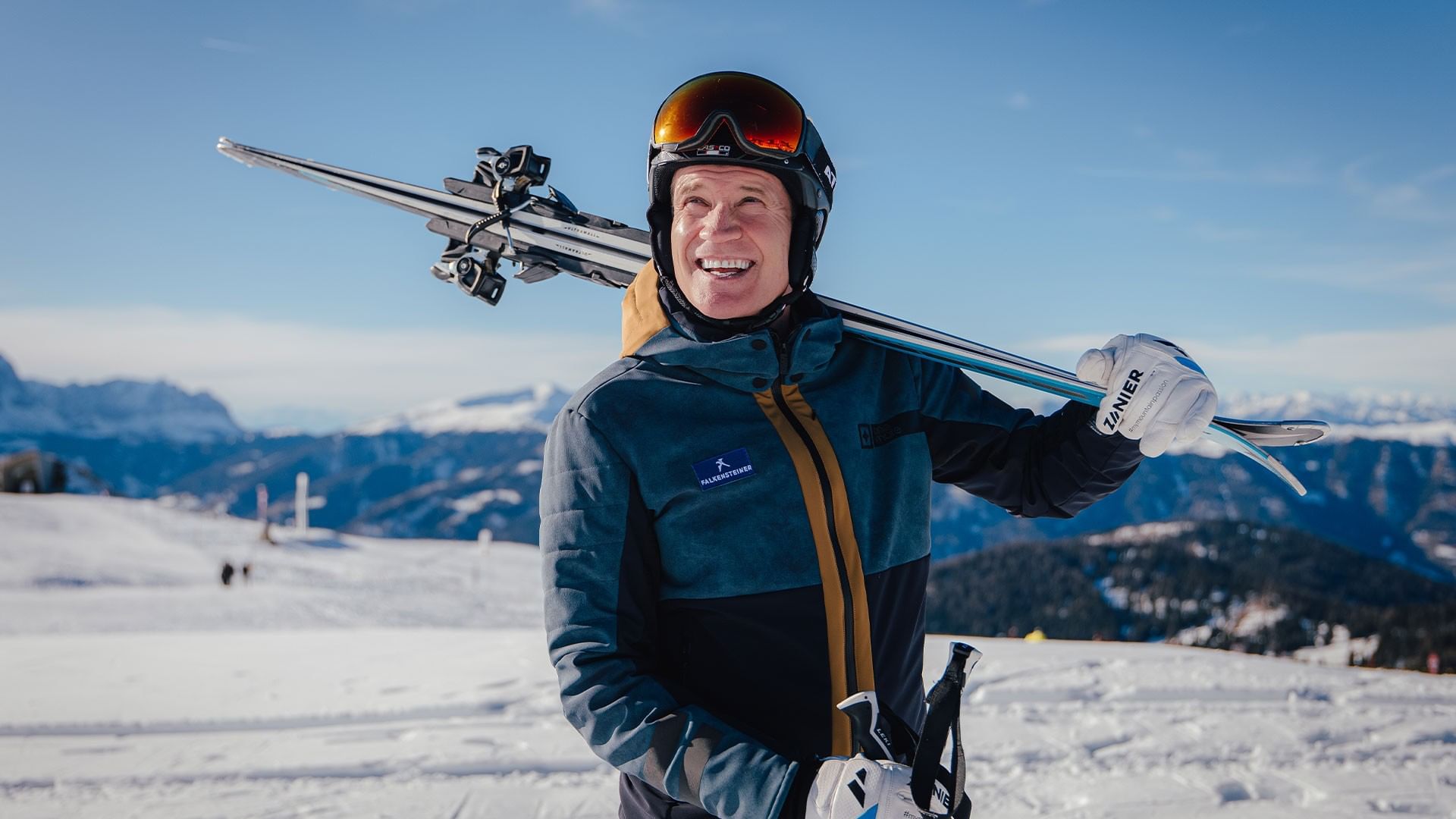 Lächelnder Mann in Skiausrüstung und mit Skiern auf den Schultern am Berg bei Falkensteiner Group