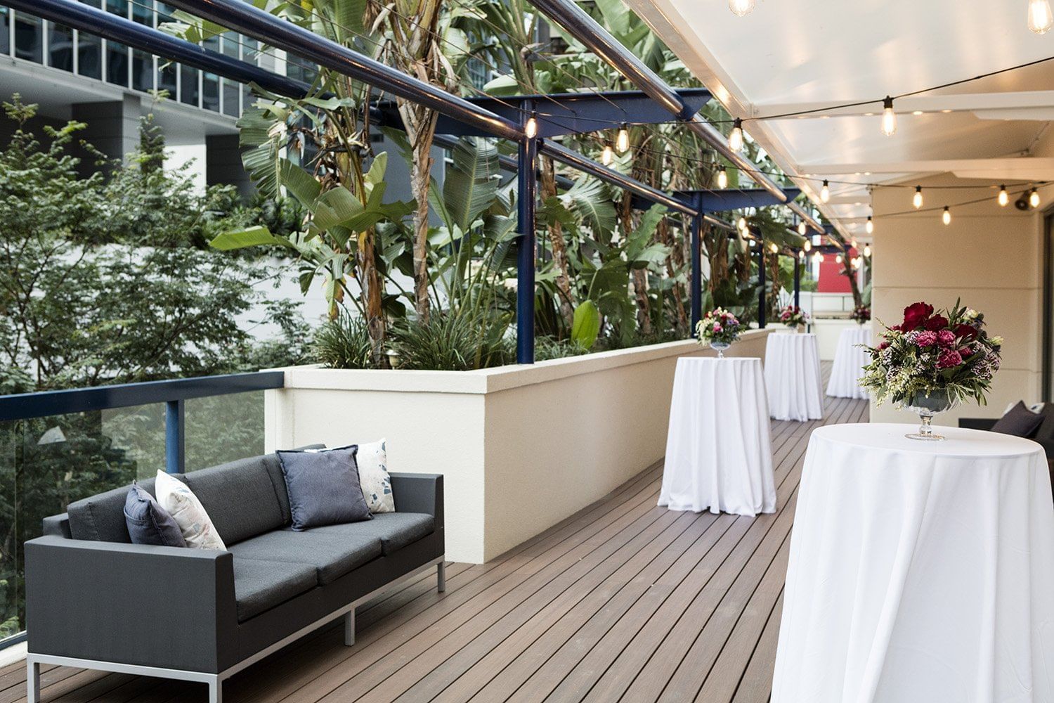 Outdoor timber balcony deck with a dark sofa, cocktail tables, lush greenery, and string lights at The Sebel Brisbane
