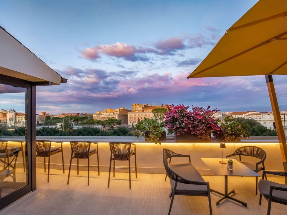 Rooftop Terrazza Costanza at The Guardian Hotel with stylish chairs, a table, vibrant flowers, and a city view at sunset