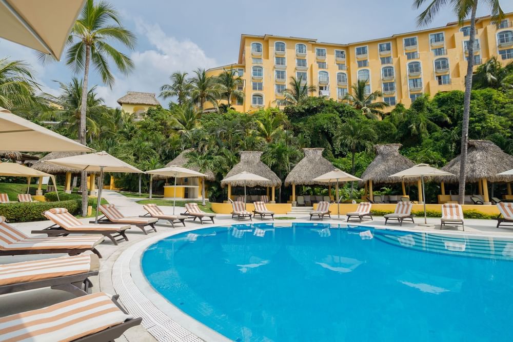 Scenic pool area with yellow hotel buildings, palm trees, and thatched roof huts at Quinta Real Acapulco