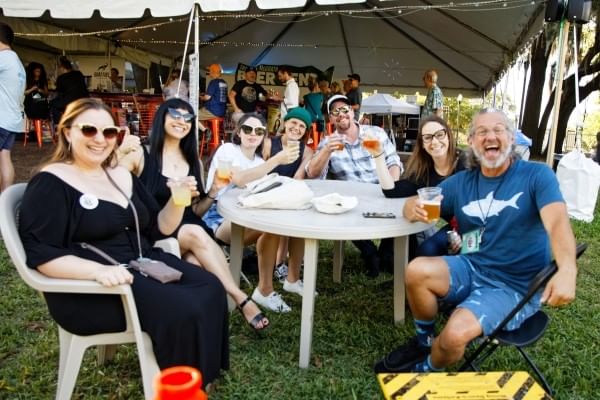 A group of people smile at the camera, sitting around an outdoor table with cups of beer in hand.