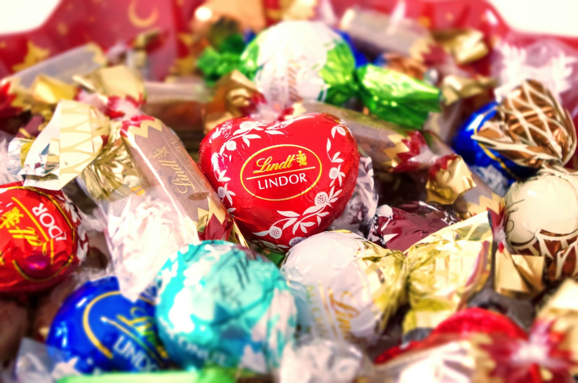 A close-up shot of individually wrapped Lindt Lindor chocolate truffles in various colorful wrappers, including red, blue, gold, green, and white. A prominent red heart-shaped truffle with the "Lindt Lindor" logo is in the center.