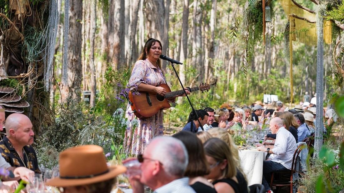 Female musician performing with acoustic guitar for seated guests at outdoor forest dining event for Pair'd Margaret River Region 2026