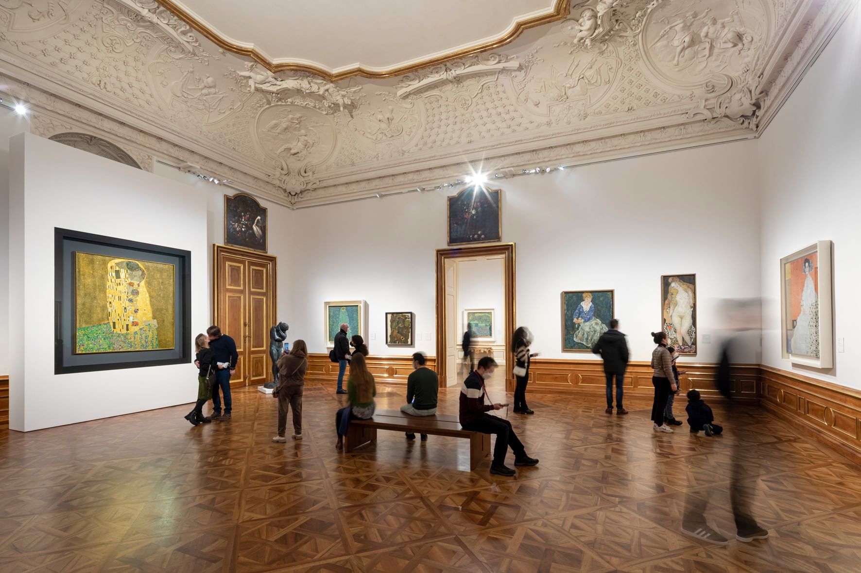 Ausstellungsraum im Belvedere in Wien mit Besuchern vor Gemälden von Gustav Klimt, darunter „Der Kuss“, in einem prunkvollen historischen Saal.  EN: