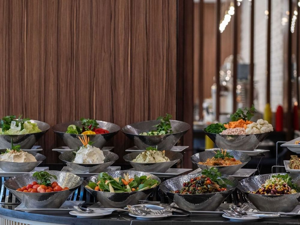 Salad bar featuring bowls of fresh greens and dips by a wood wall surrounding a buffet area at Warwick Al Jubail