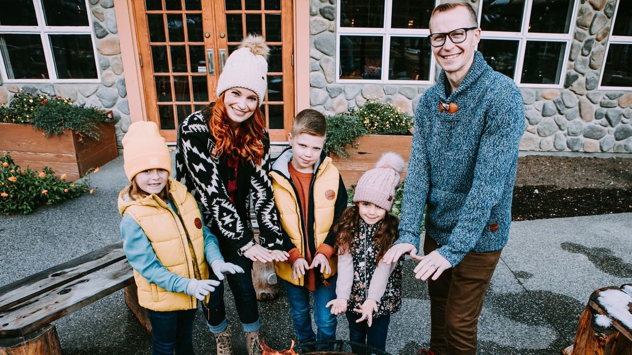 A family of five dresses for a winter vacation in Canmore, standing outside warming their hands on a fire.