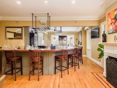 Wooden barstools by a curved bar, under hanging wine glasses near a fireplace, and framed art at Simsbury 1820 House