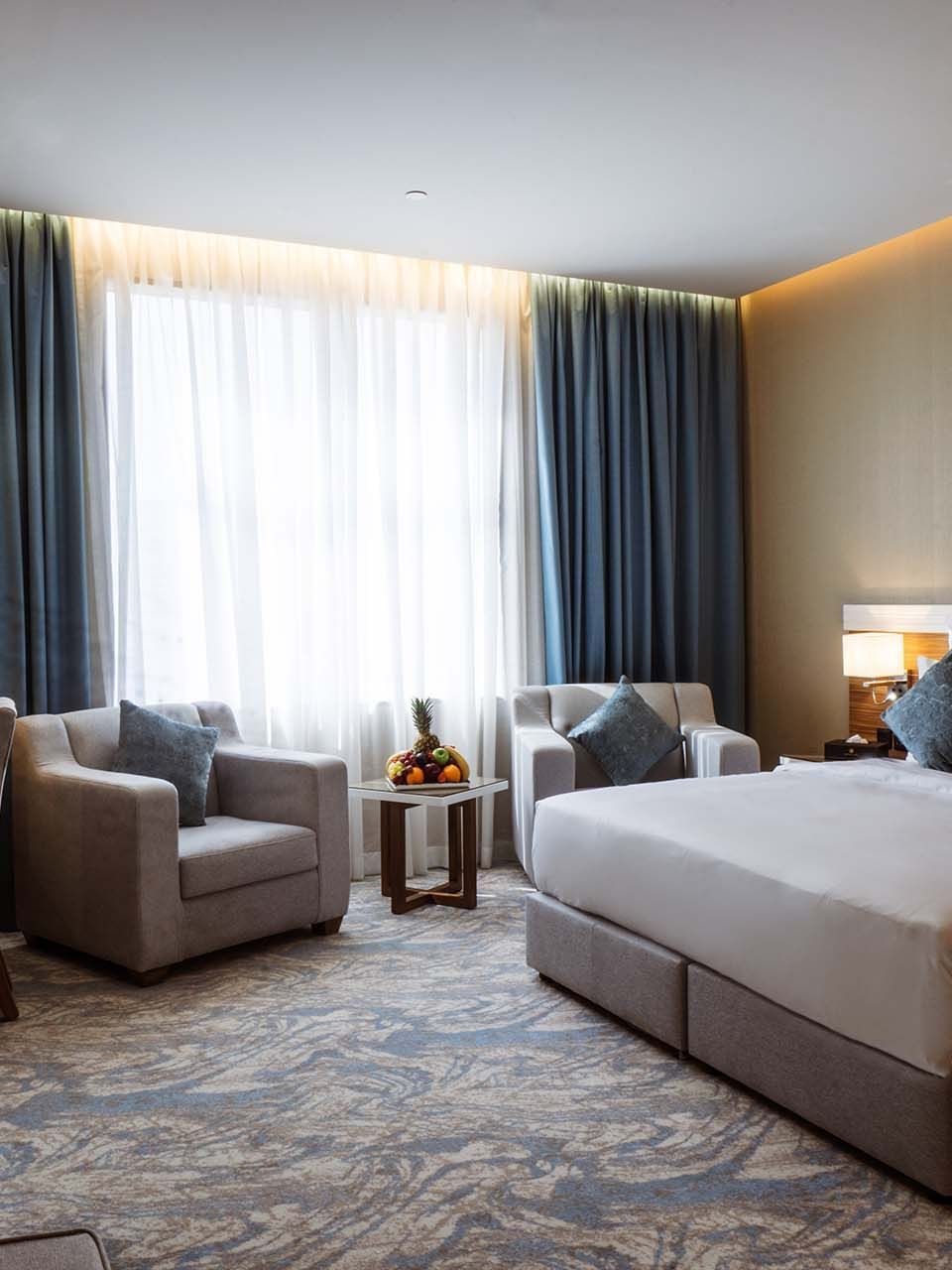 Large bed with armchairs by a fruit tray under a large window in Standard King Room at Cantonal Hotel by Warwick Riyadh