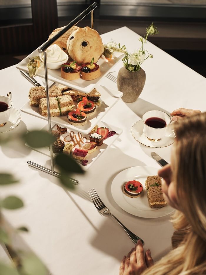 Woman at a dining table with a three-tier dessert stand and tea, looking at her plate.