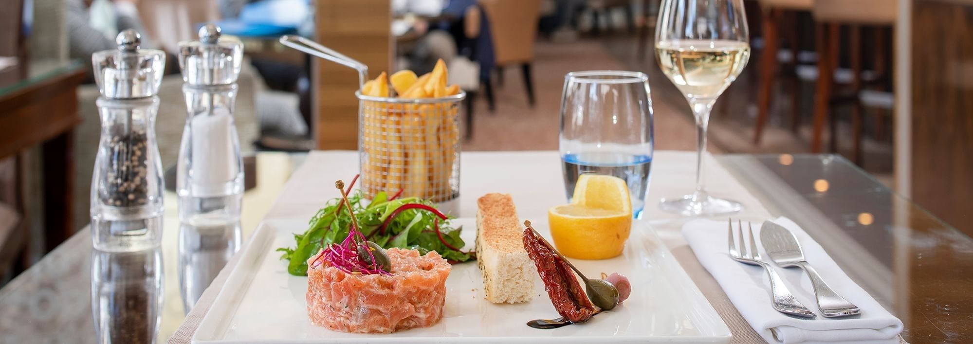 Plated salmon tartare with mixed greens, fries, toast and dried tomato beside a glass of white wine at Téséo Restaurant & Bar