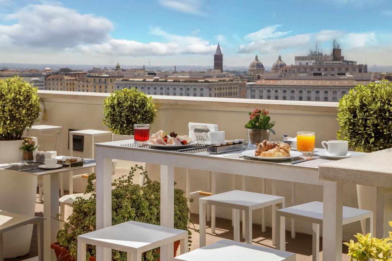 Breakfast buffet with fresh fruit, pastries, and juices in a modern rooftop lounge with outdoor seating and city skyline views.