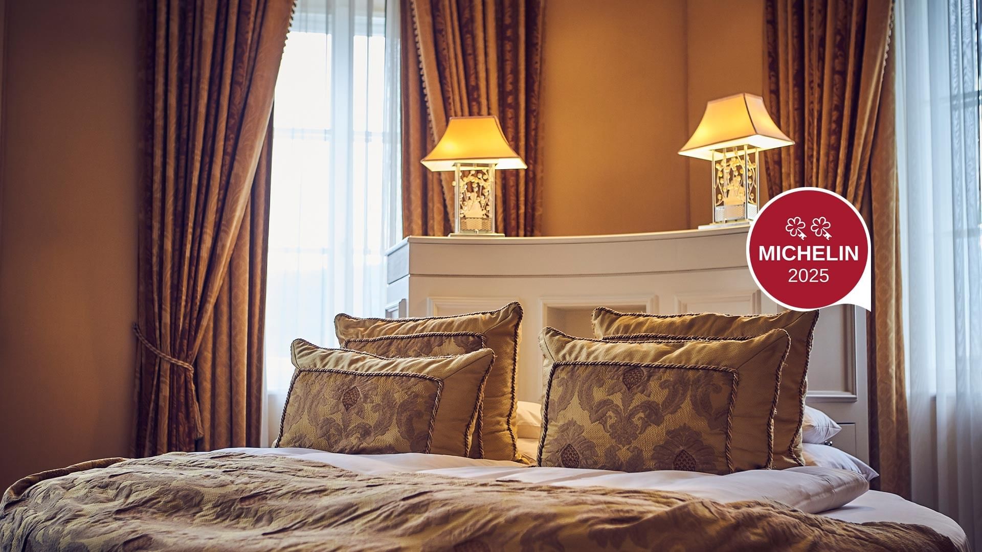 Letto elegante con cuscini decorativi e lampade da tavolo, finestra con tende pesanti e logo Michelin 2025.