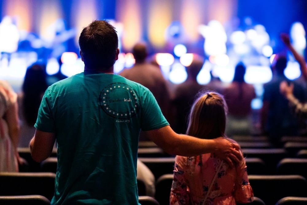 A dad in a blue t-shirt rests his hand on his daughter’s shoulder as they face a stage at a concert.