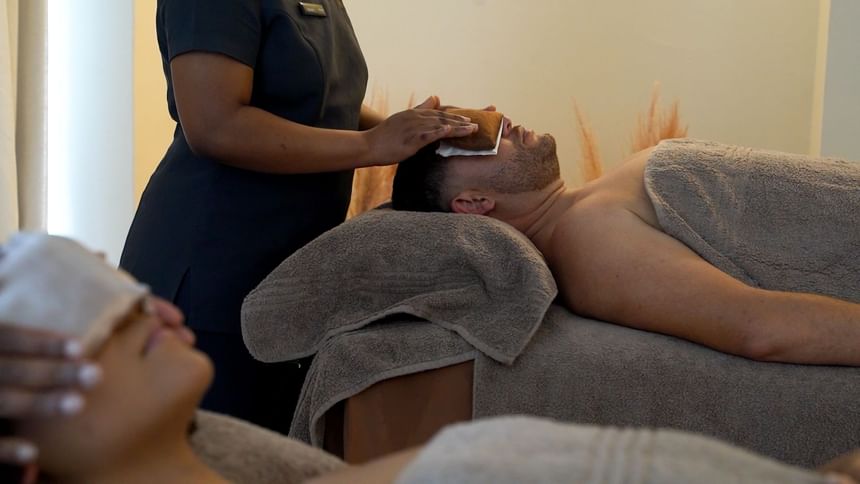 Two people receiving facials from a spa technician in a room with beige walls and white curtains at Pepperclub Hotel