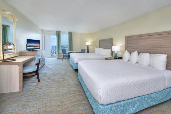 A wide-angle view of a hotel room with two queen beds, a desk, television, and balcony.