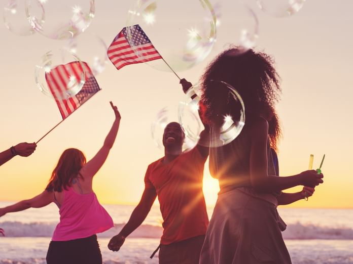 Grupo de personas celebrando con banderas estadounidenses y burbujas en la playa al atardecer