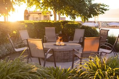 Chairs surround a fire pit in a garden with a tree and a building in the background.
