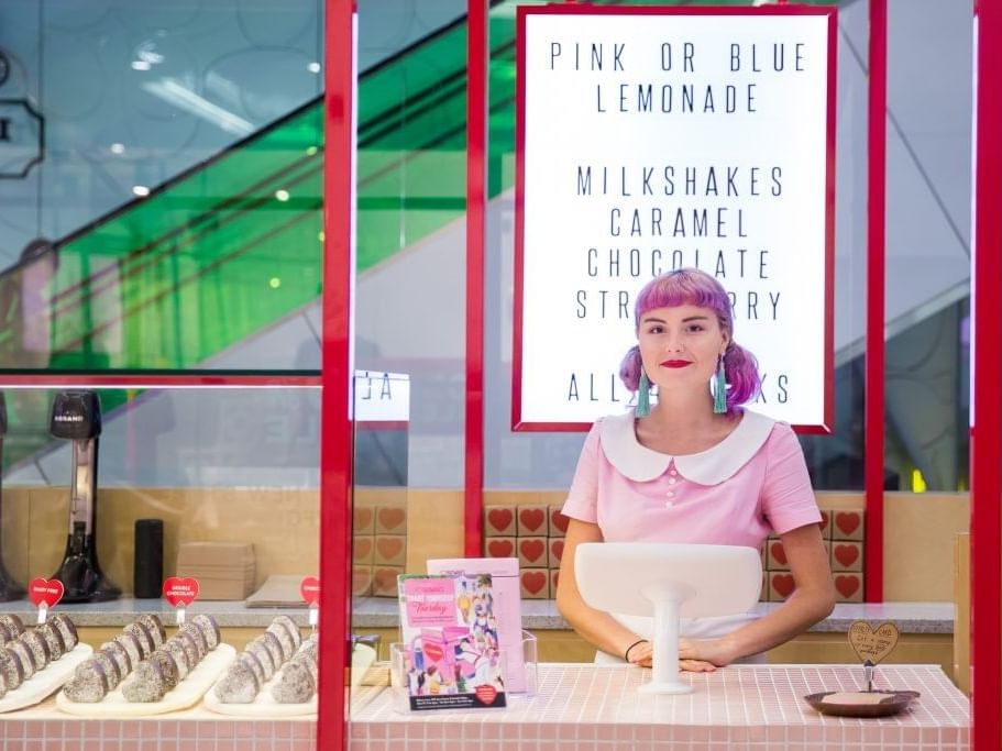 Woman with pink hair at a bright, retro-styled milkshake counter, ready to serve drinks and lemonade at The Sebel Brisbane