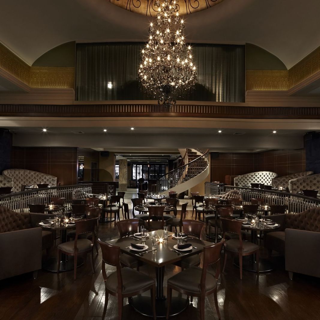 A luxurious dining room with chandeliers, dark wooden tables, upholstered booths, and a central spiral staircase.
