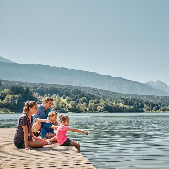 Familie genießt den Ausblick auf einen See mit Bergen im Hintergrund, während sie auf einer Holzplattform sitzt.
