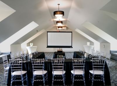 A meeting room setup at The White House Hotel with a screen, black tables, and clear chairs in a bright attic space