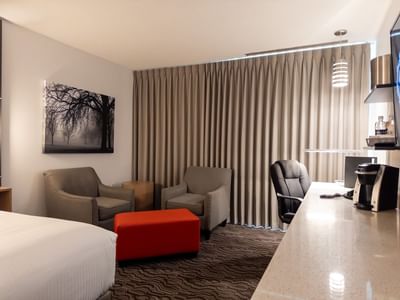 A hotel room with a bed, two armchairs, a red ottoman, and a desk from Carriage House Hotel & Conference Centre