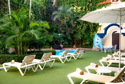 Área de piscina con camastros blancos, sombrilla y exuberante vegetación tropical en Los Arcos Suites