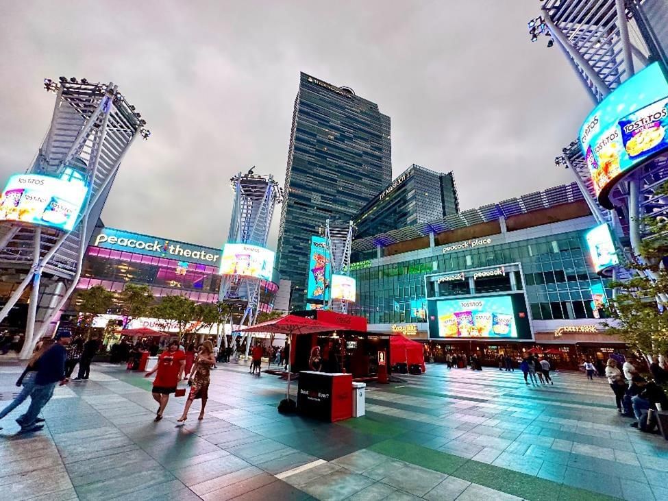 A wide-angle shot of a bustling city square with a large digital billboard and several tall buildings in the background.
