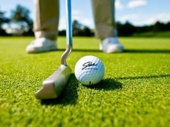 Person's legs and golf club poised to hit a golf ball on green grass with blue sky in background.