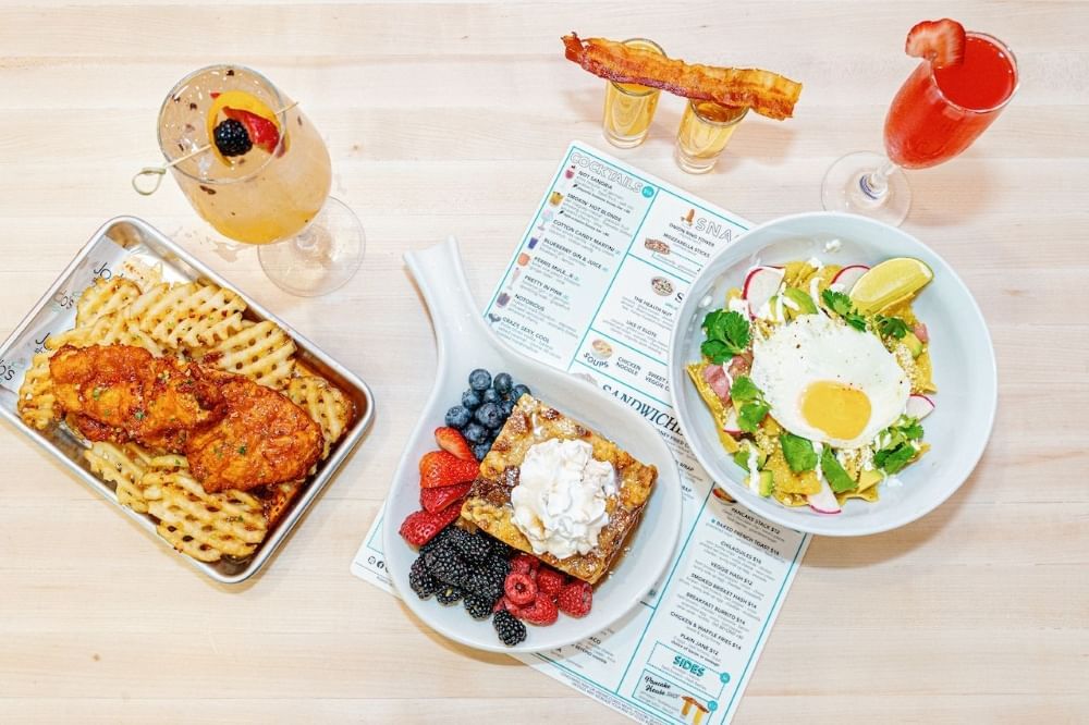 An aerial shot of various brunch dishes and cocktails on top of a menu with a light wood background.