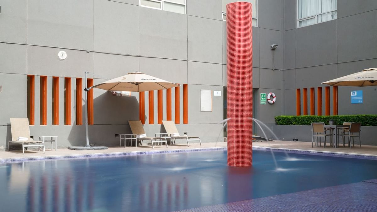 Área de piscina moderna con fuente alta de mosaico rojo y tumbonas en Real Inn Torreón