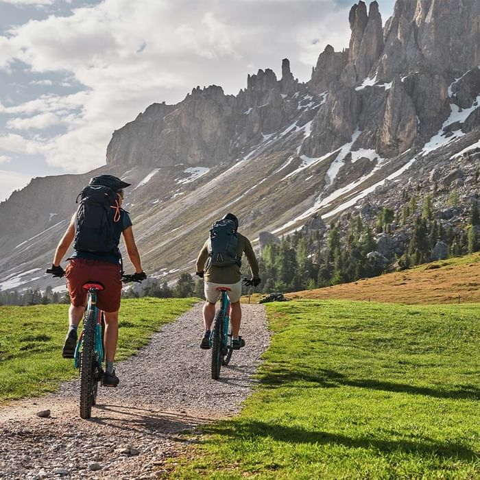 Due ciclisti con zaini percorrono un sentiero in una zona montuosa con neve e vegetazione.