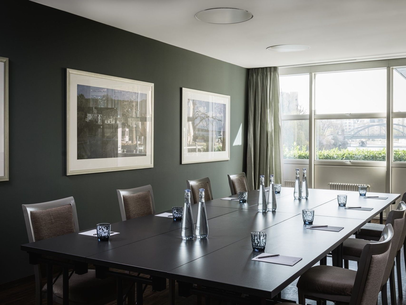 Konferenzraum mit langem Tisch, Stühlen, gerahmten Bildern und großen Fenstern mit Blick auf die Stadt.