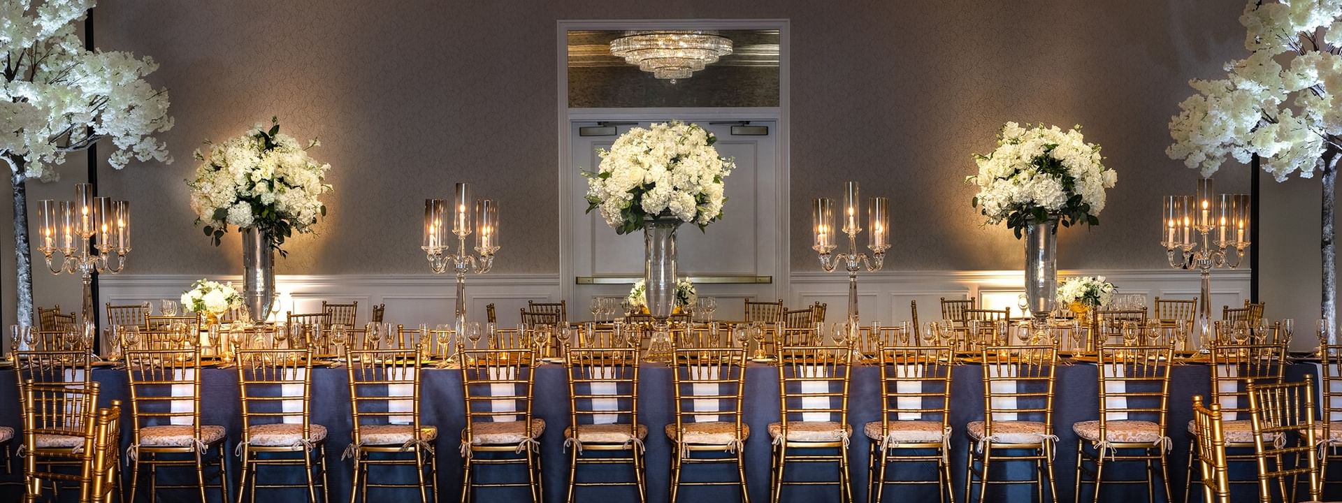 Long dining table by gold chairs under white floral towers in Turtle Creek Ballroom at Warwick Melrose Dallas