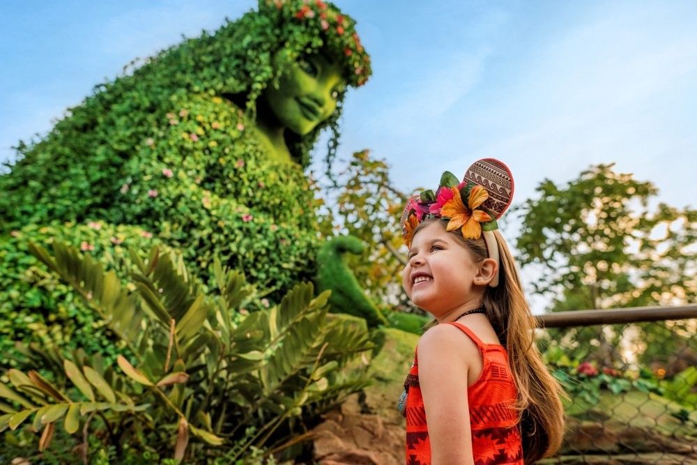 A toddler girl in a Moana costume stands in front of a topiary figure of a woman with an outstretched hand.