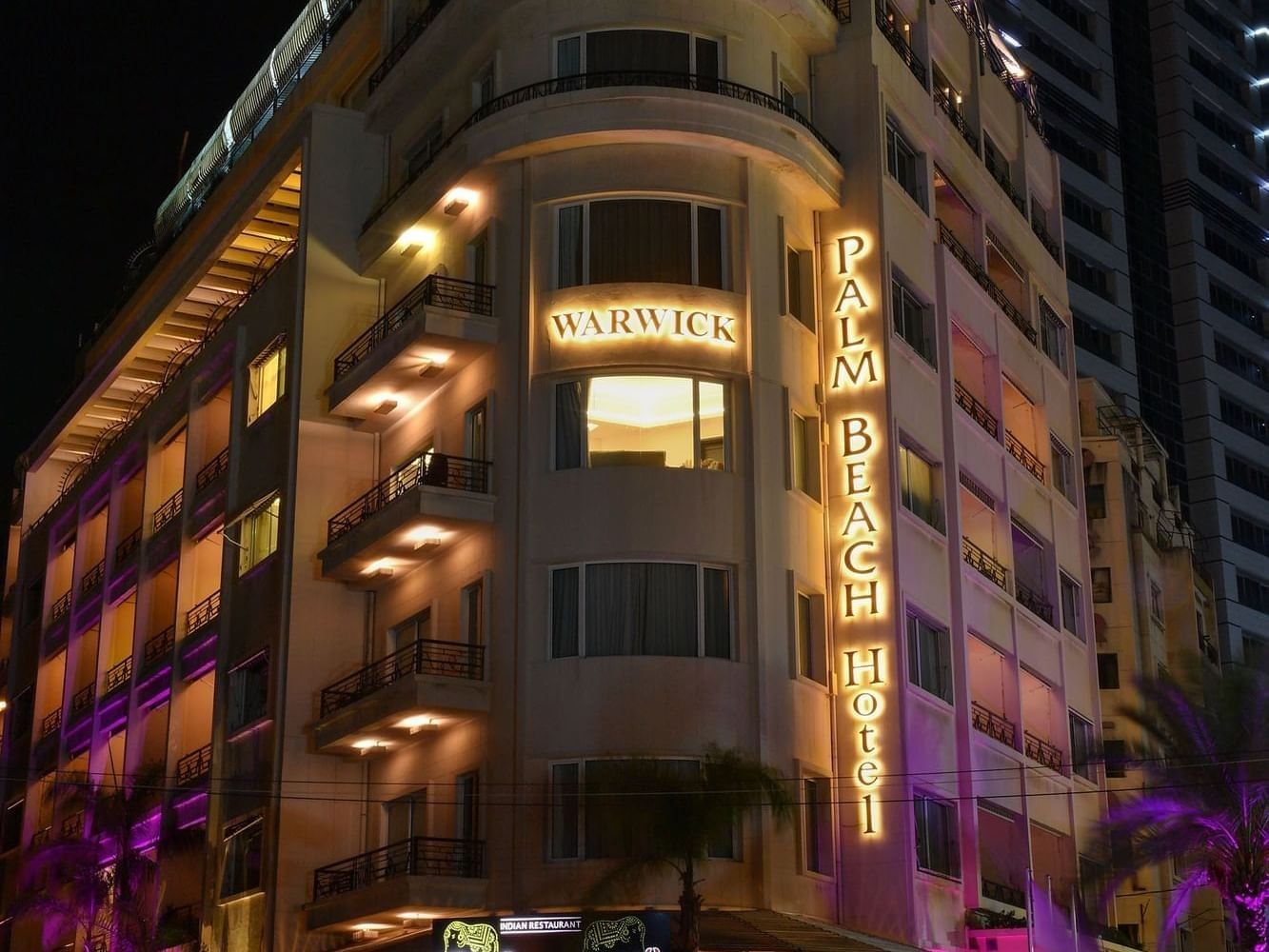 Exterior of Warwick Palm Beach Hotel at night, featuring glowing signs and purple-lit palm trees